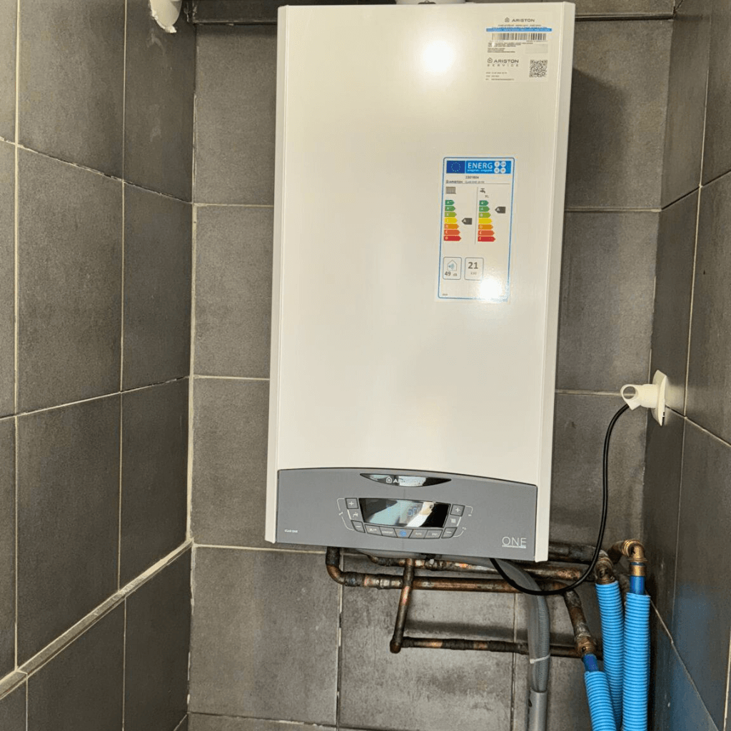 Chaudière gaz murale Ariston One installée sur un mur carrelé gris dans une buanderie, avec tuyauterie cuivre et gaines bleues, pour un chauffage performant en Hauts-de-France.